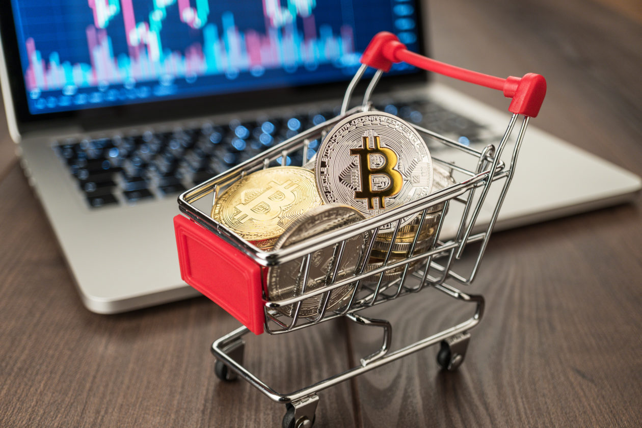 Physical models of Bitcoin in a miniature shopping cart in front of a laptop displaying price graphs