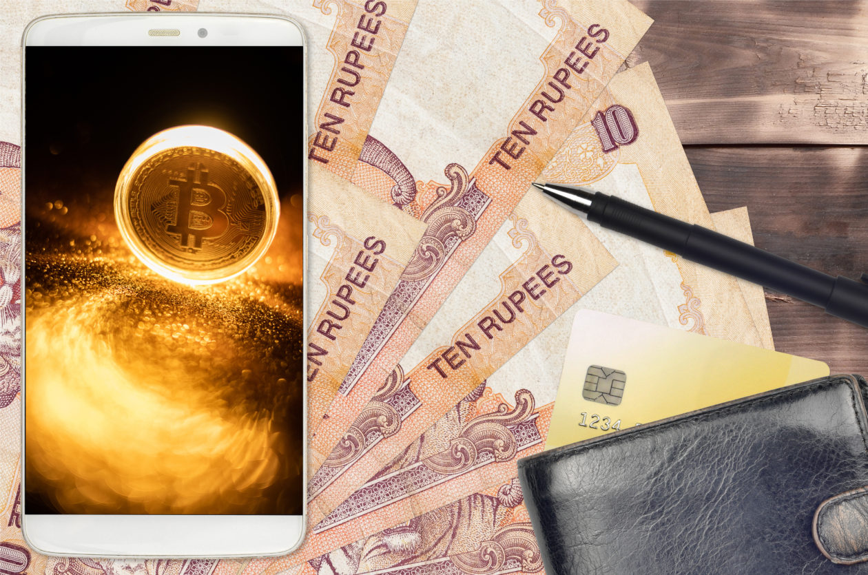 Bitcoin displayed on phone screen on top of rupees laid out on desk with pen and wallet