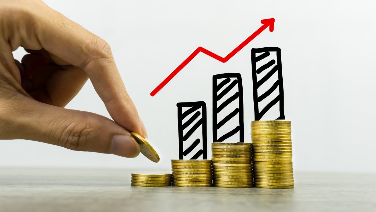 Long-term investment or making money with the right concepts. A business man putting coin on stack of coins on a table with growing graph on coins pile. Depicts a standing and stable investment.
