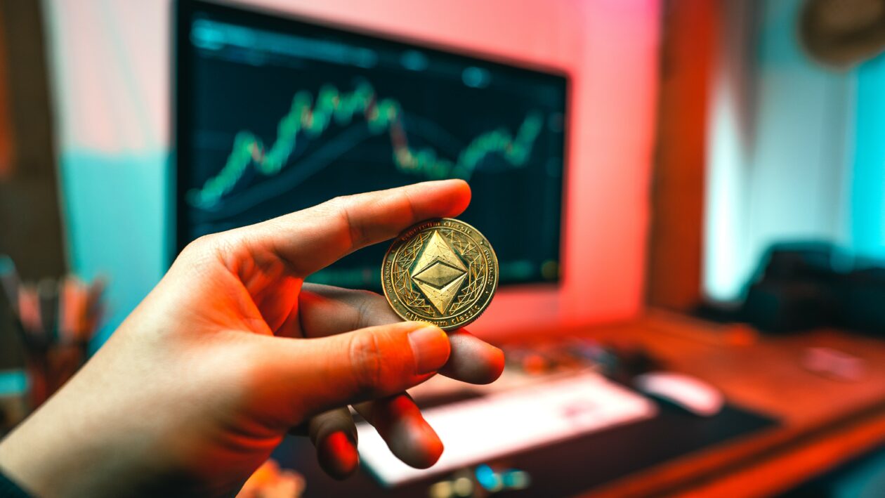 A man's hand holds an etherium coin at a computer table, Markets: Etherium Merge complete, ETH and linked tokens lift market