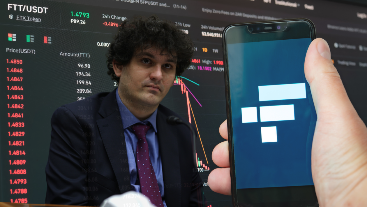 FTX CEO SBF sitting behind a smart phone holding an app displaying FTX logo