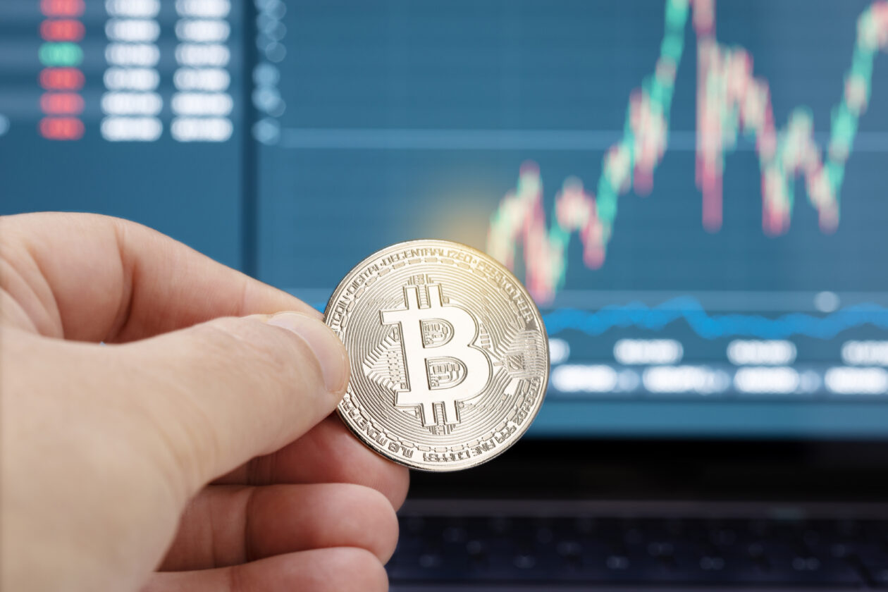Hand holding a bitcoin coin and chart on laptop display at background.